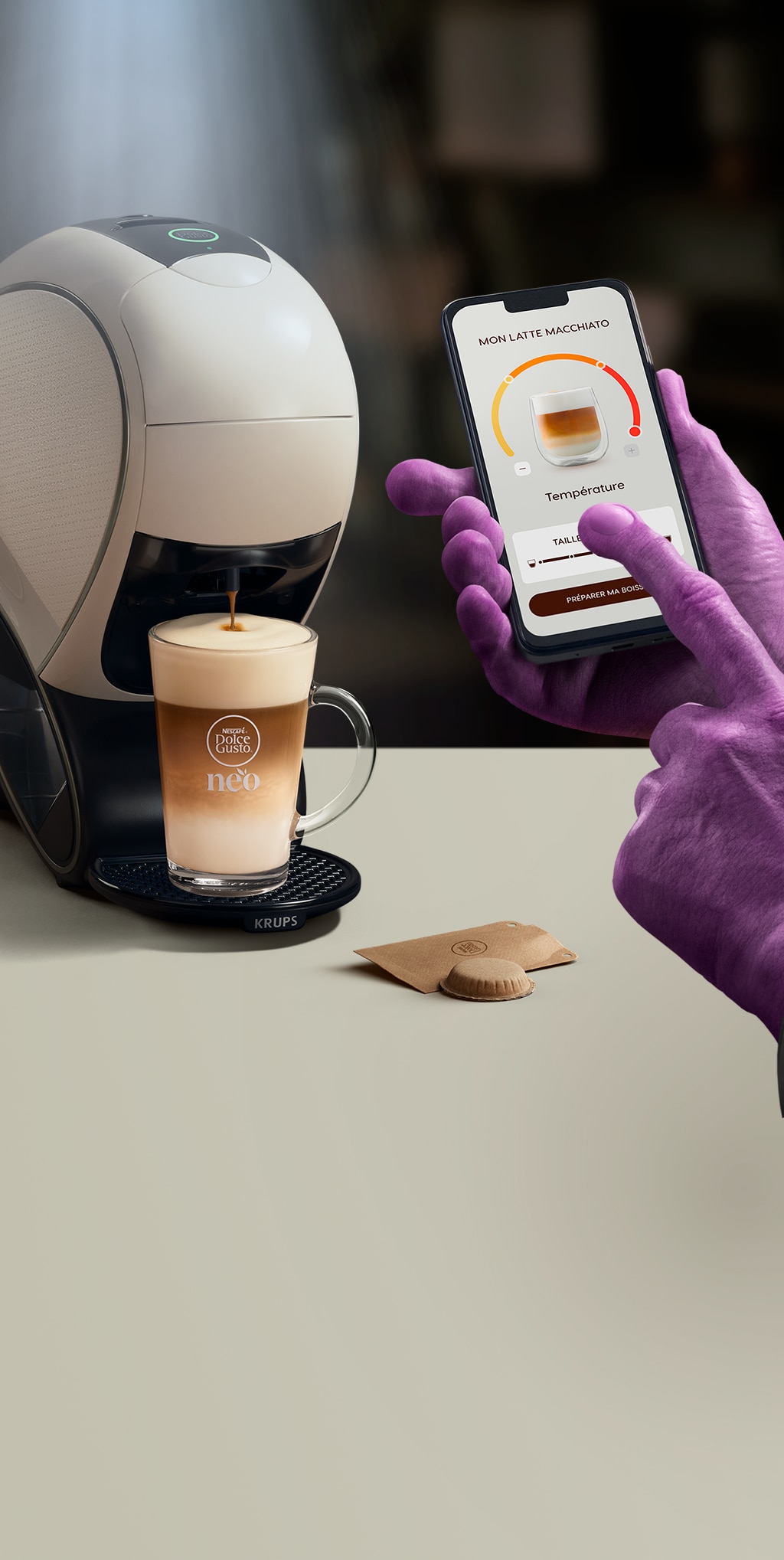 Main d'un extraterrestre utilisant l'application NESCAFE Dolce Gusto pour personnaliser son café. En arrière-plan, la machine NEO, des boîtes de dosettes et des boissons.