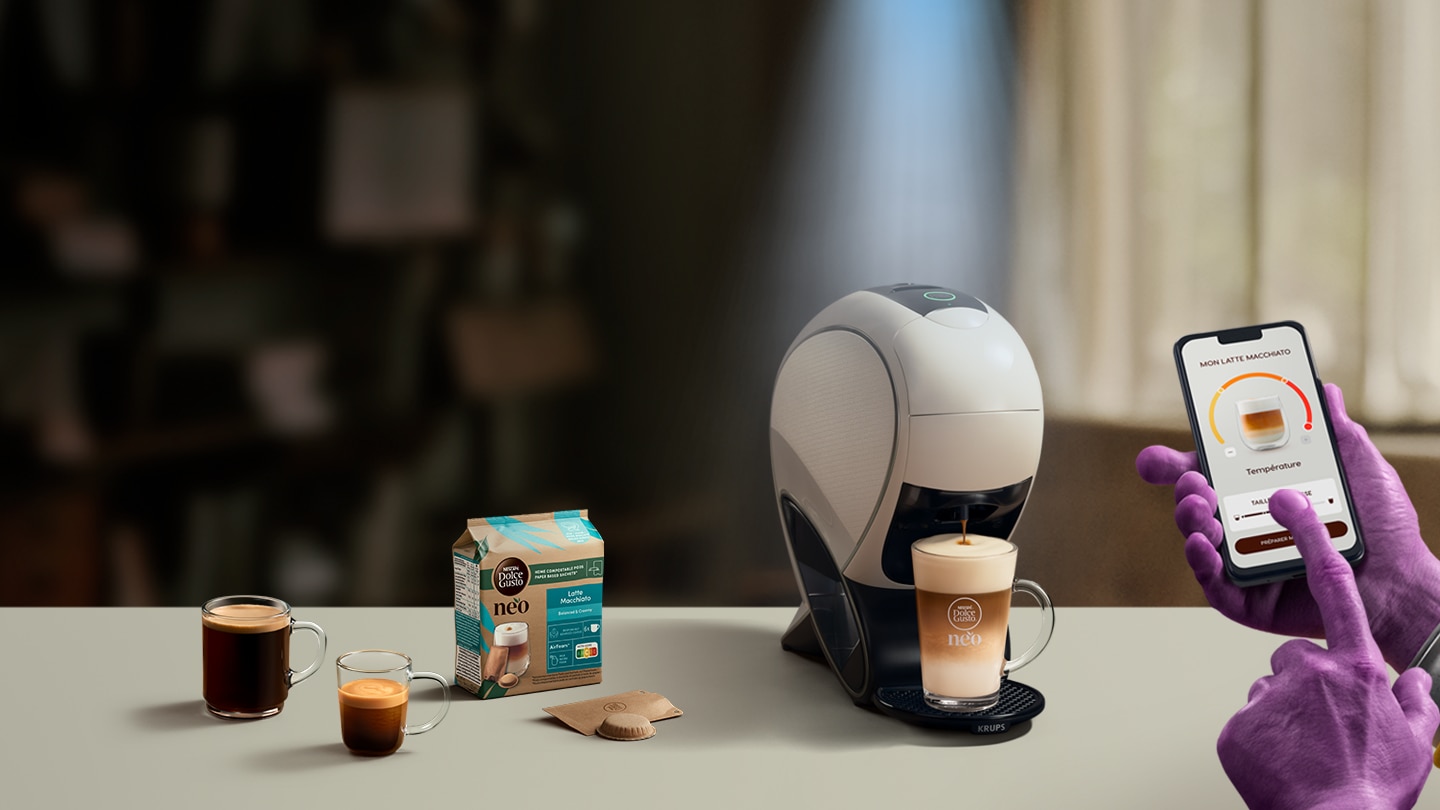 Main d'un extraterrestre utilisant l'application NESCAFE Dolce Gusto pour personnaliser son café. En arrière-plan, la machine NEO, des boîtes de dosettes et des boissons.
