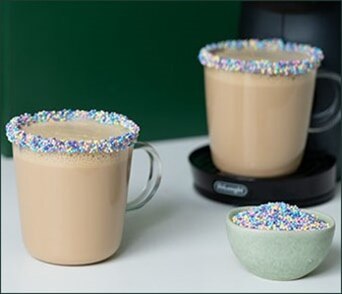 Deux mugs de café crémeux aux bords délicatement décorés de vermicelles colorés, accompagnés d’un petit bol de vermicelles pour une touche gourmande personnalisée.